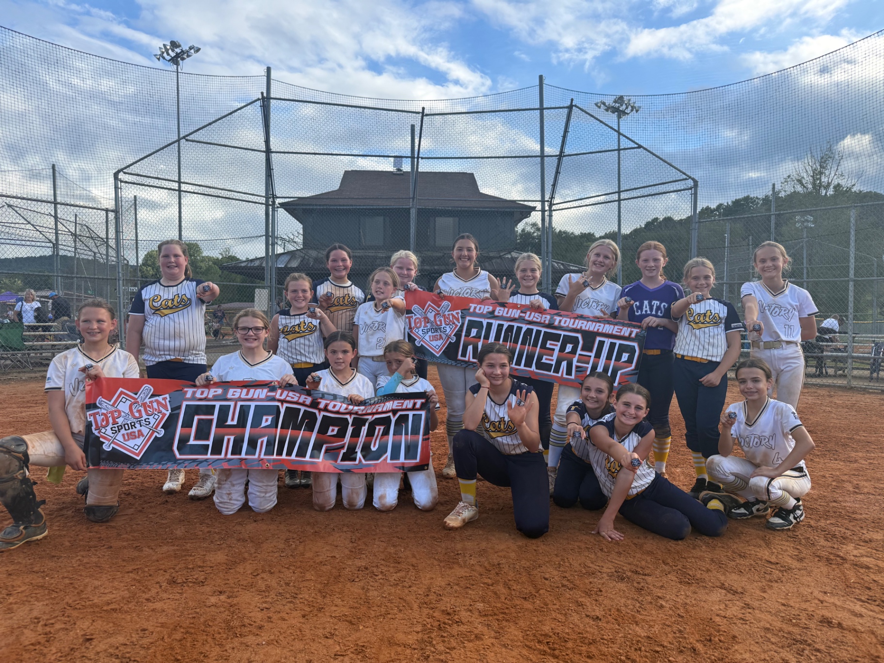 Victory softball players and fellow tournament champions posing together on the field with championship banners and award rings.
