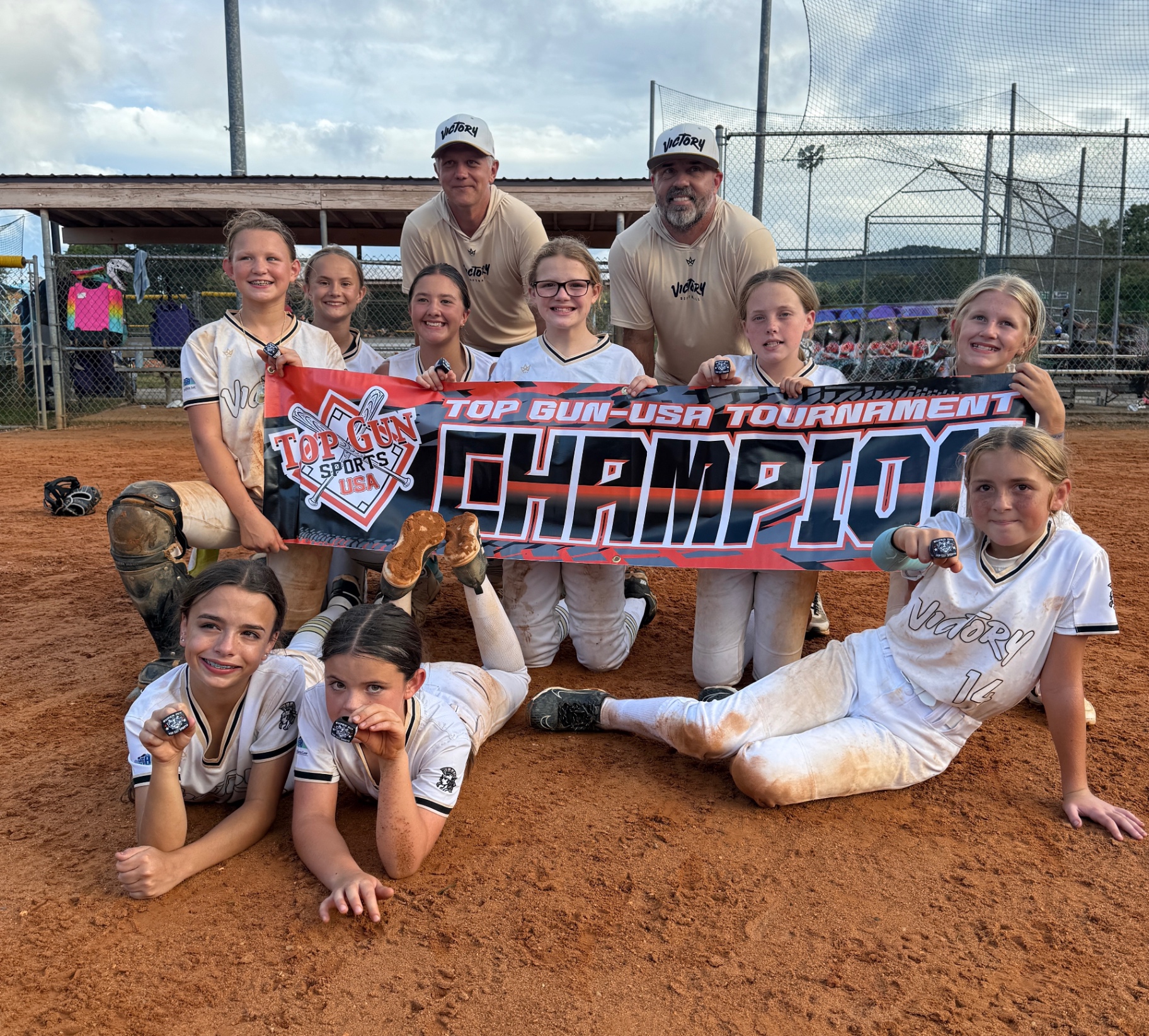 Victory softball players and coaches posing on the field with a Top Gun USA Tournament Champions banner and award rings.