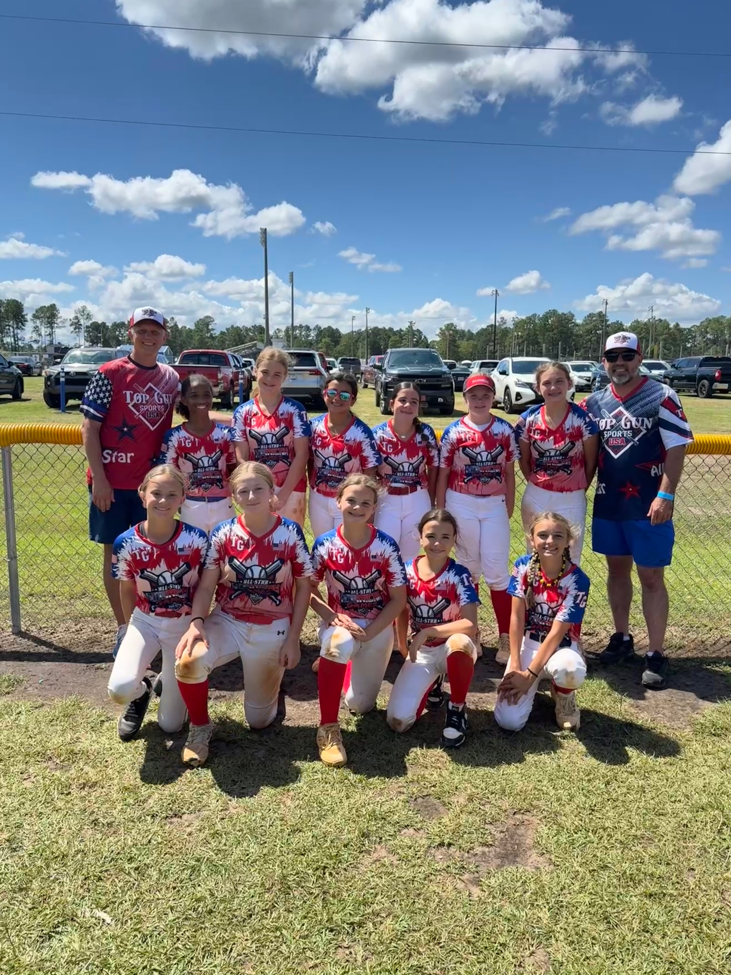The Top Gun All-Stars team and coaches posing together in front of the outfield fence on a sunny day.