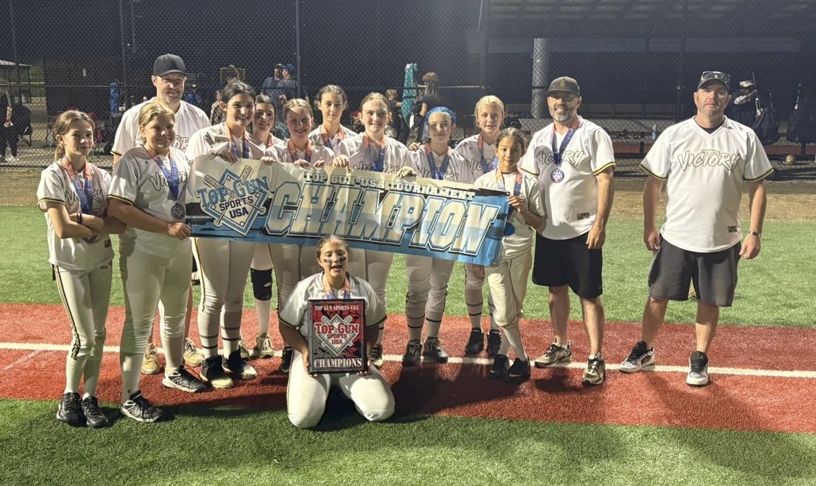 Softball team and coaches gathered on turf field holding a victory champion banner and award plaque.