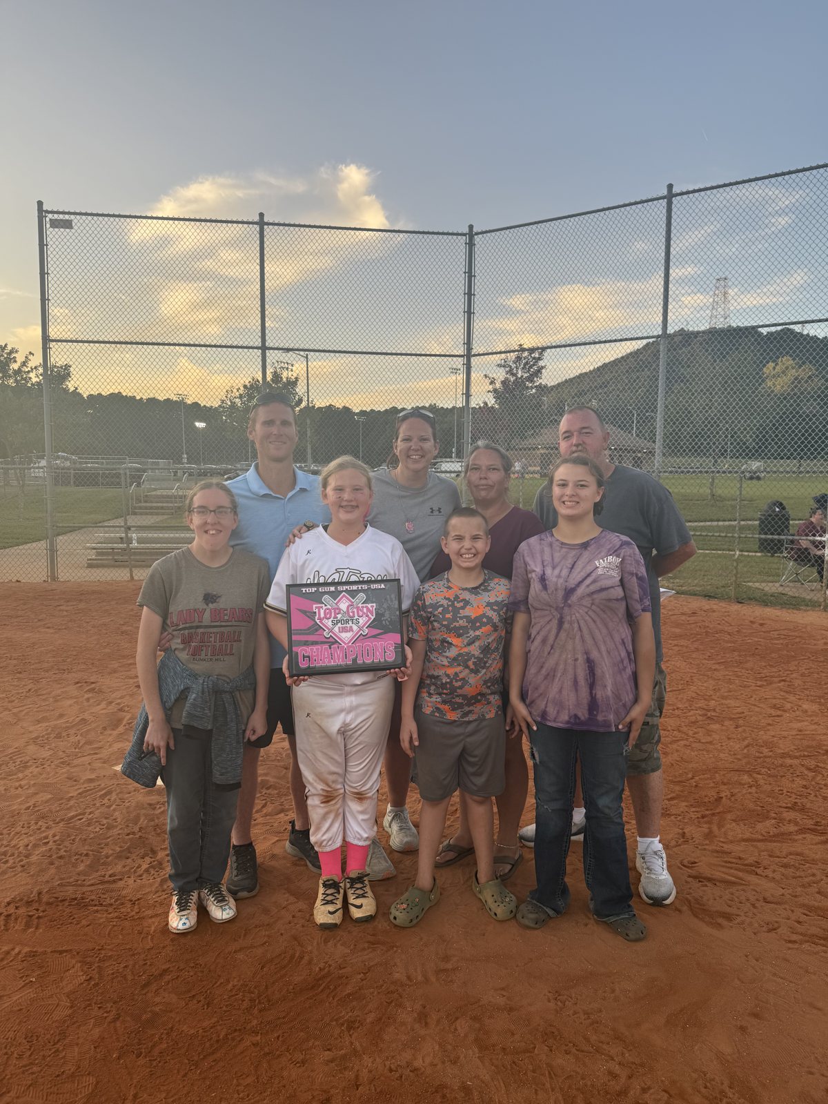 A group of players and adults pose on the dirt field holding a pink championship plaque.