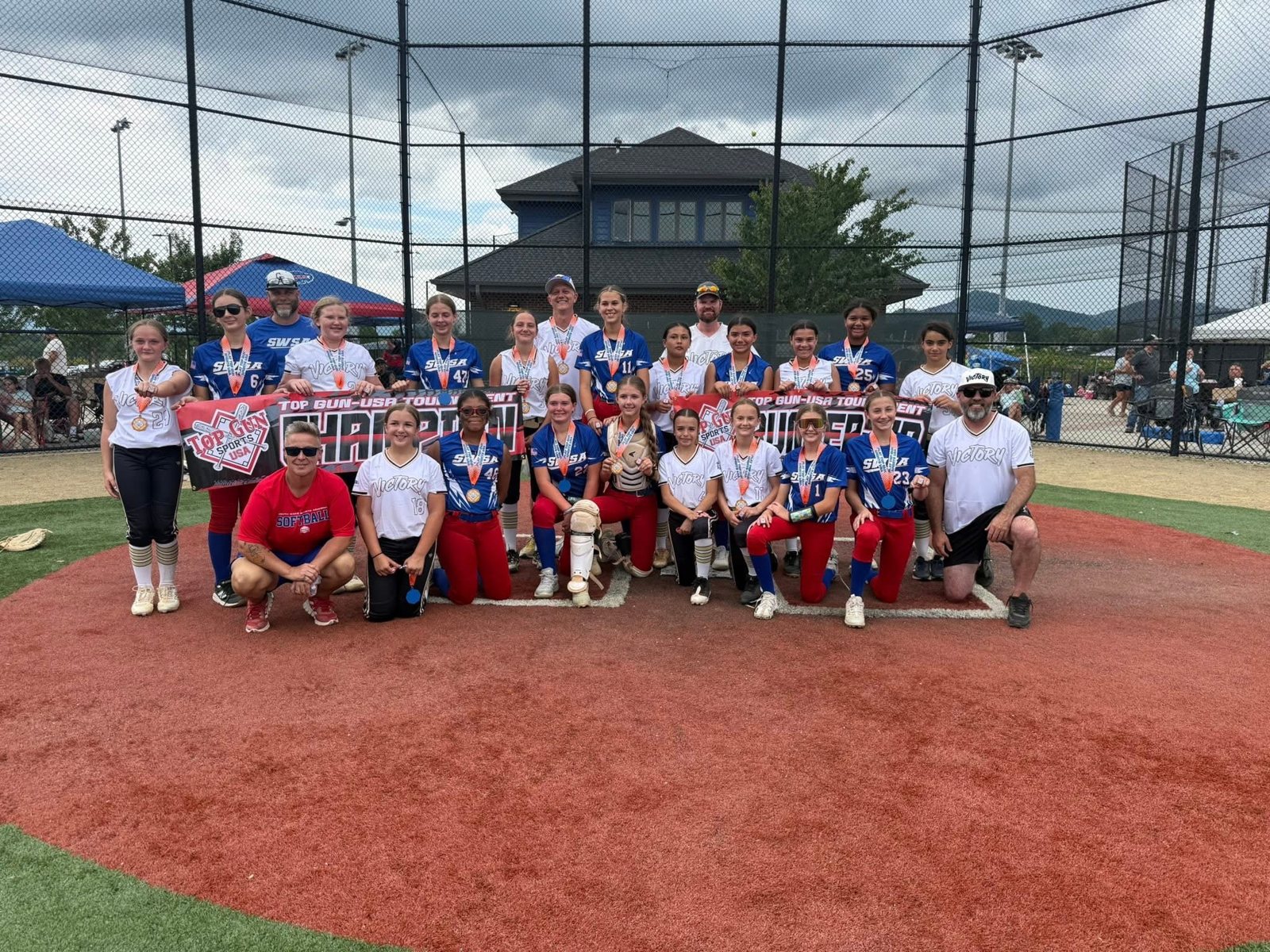A softball team gathered on a dirt field posing for a team portrait holding a banner and awards.