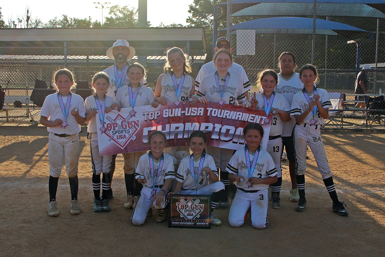 A youth softball team and coaches posing together behind a championship banner and plaque on the field.